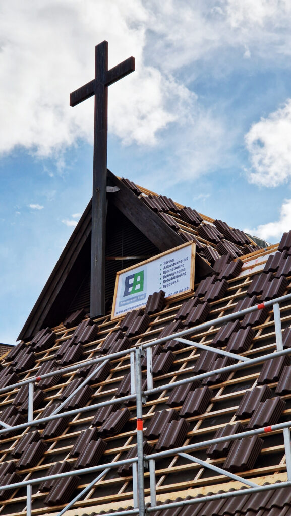 Bilde viser et tak under konstruksjon eller renovering. Taket er dekket av mørkebrune takstein som er montert på et stillasystem. I midten av bildet rager et stort kors opp mot himmelen, omgitt av de brune taksteinene. Nederst i bildet er det synlig et skilt med kontaktinformasjon for et byggefirma.