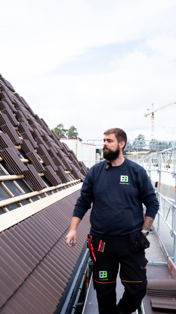Bilde viser en arbeider fra byggefirmaet som står og ser ut over byggeplassen. Arbeideren har på seg en blå genser med logoen til firmaet og bærer også et belte med verktøy. I bakgrunnen er det synlig mørkebrune takstein som er arrangert i rader, og i det fjerne er det også synlig et krankonstruksjon.