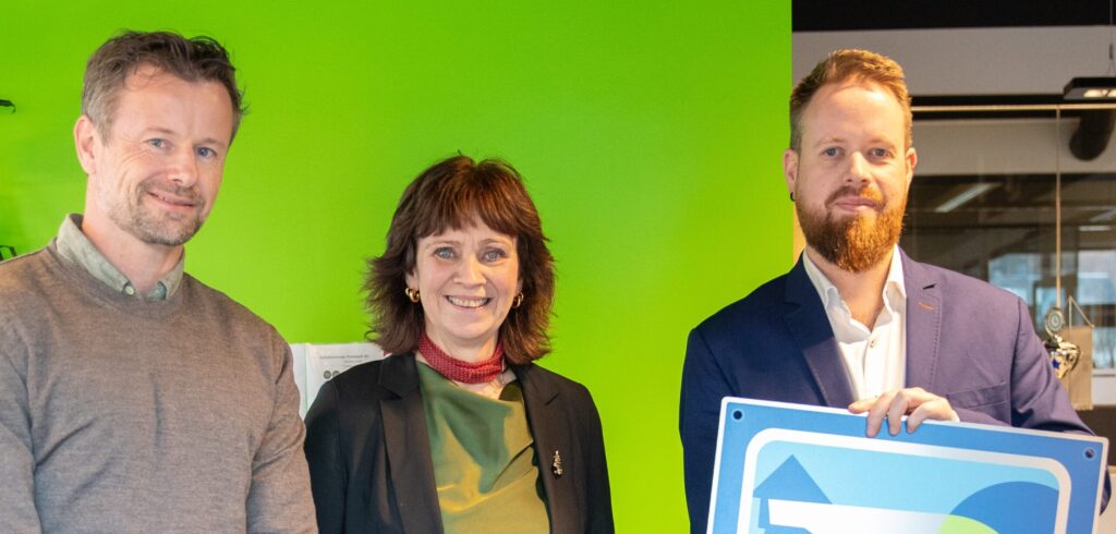 Bildet viser tre personer som står foran et grønt lerret. Til venstre ser vi en mann i moden alder med grått hår og skjegg, kledd i en grå genser. I midten ser vi en kvinne i moden alder med kort mørkt hår, kledd i en svart jakke over et rød- og hvitmønstret topp. Til høyre ser vi en yngre mann med skjegg, kledd i en mørkeblå dress. Mannen til høyre holder et blått skilt med et geometrisk mønster, hvor det står Miljøfyrtårn.